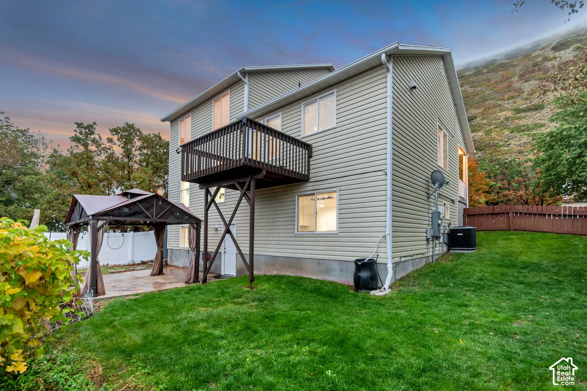 Two-story residence featuring light-colored horizontal siding, an elevated wood-framed balcony, a concrete rear patio, and a fenced yard situated against a hillside landscape