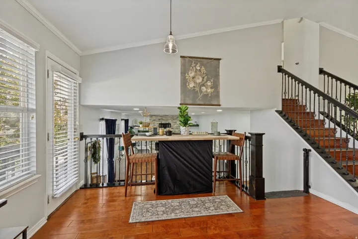 Vaulted ceiling with crown molding, wood-finish flooring, white window blinds, and dark-stained staircase with wrought iron balusters