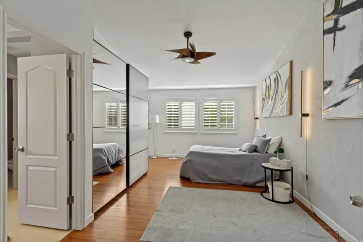 Wood-finish flooring, mirrored wardrobe doors, ceiling fan with integrated lighting, two windows with plantation shutters, and two vertical wall sconces