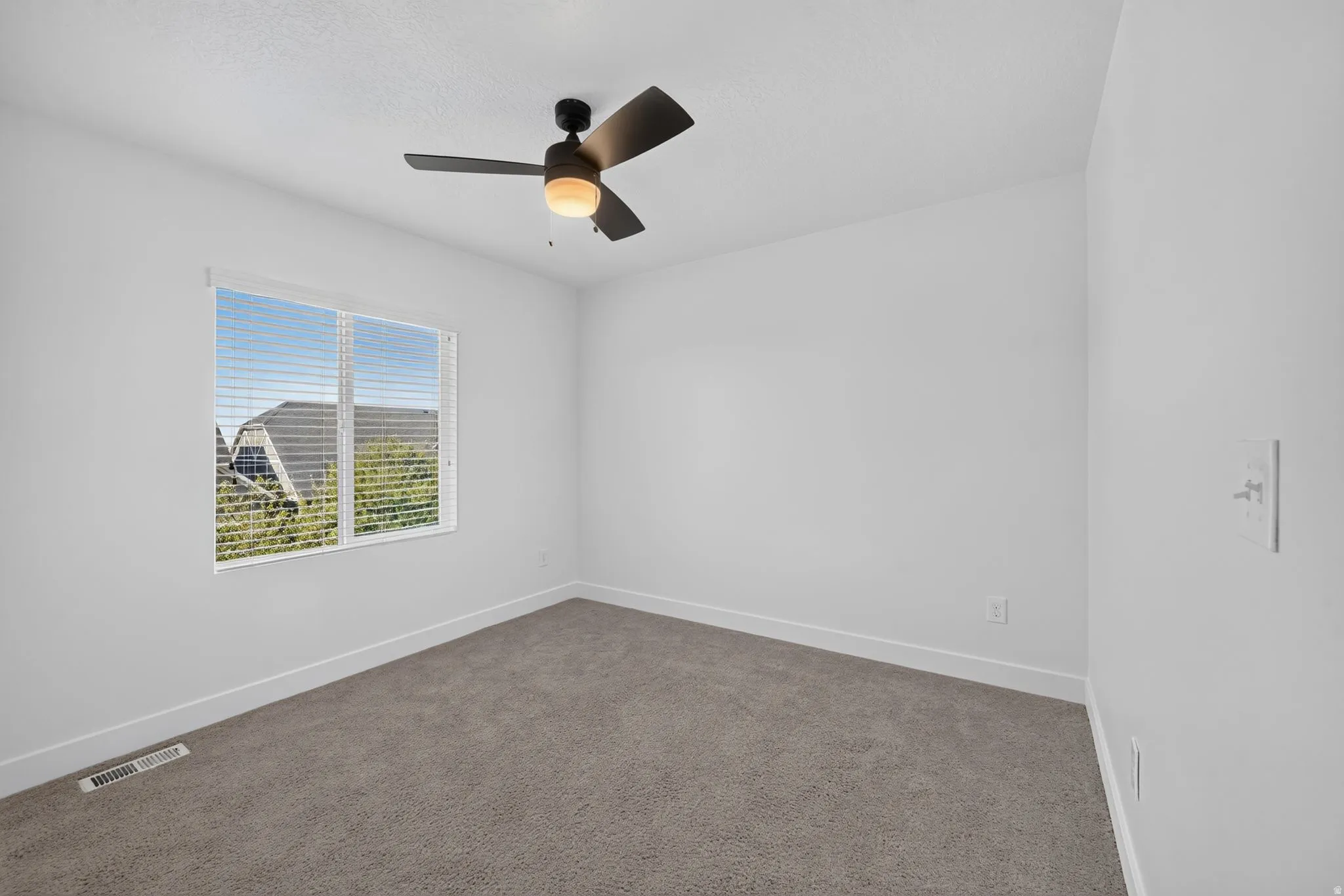 Bright secondary bedroom with carpet flooring, large double-pane window, ceiling fan with integrated light fixture, and clean white walls