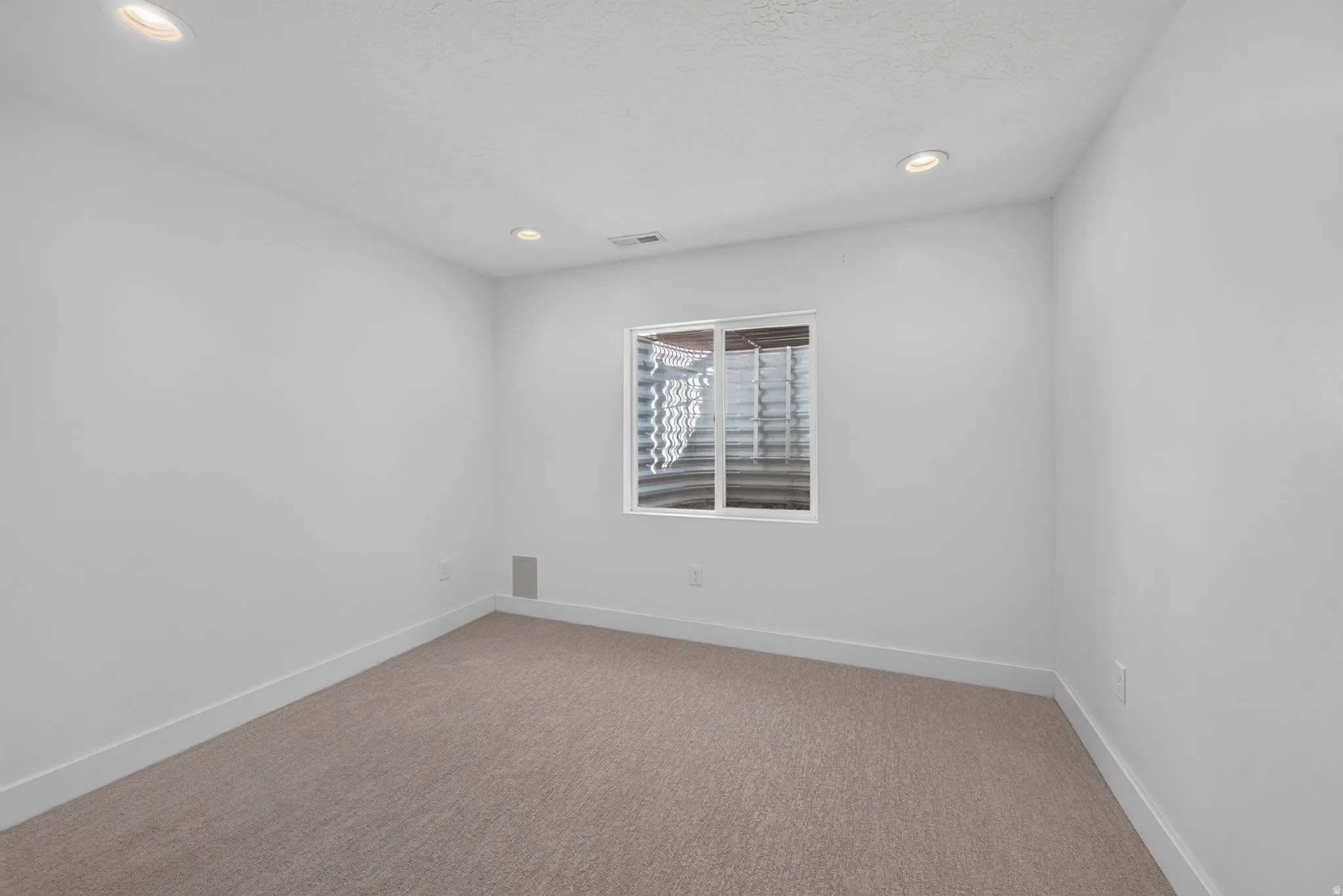 Recessed ceiling lighting, white painted walls, tan carpet flooring, and a single window