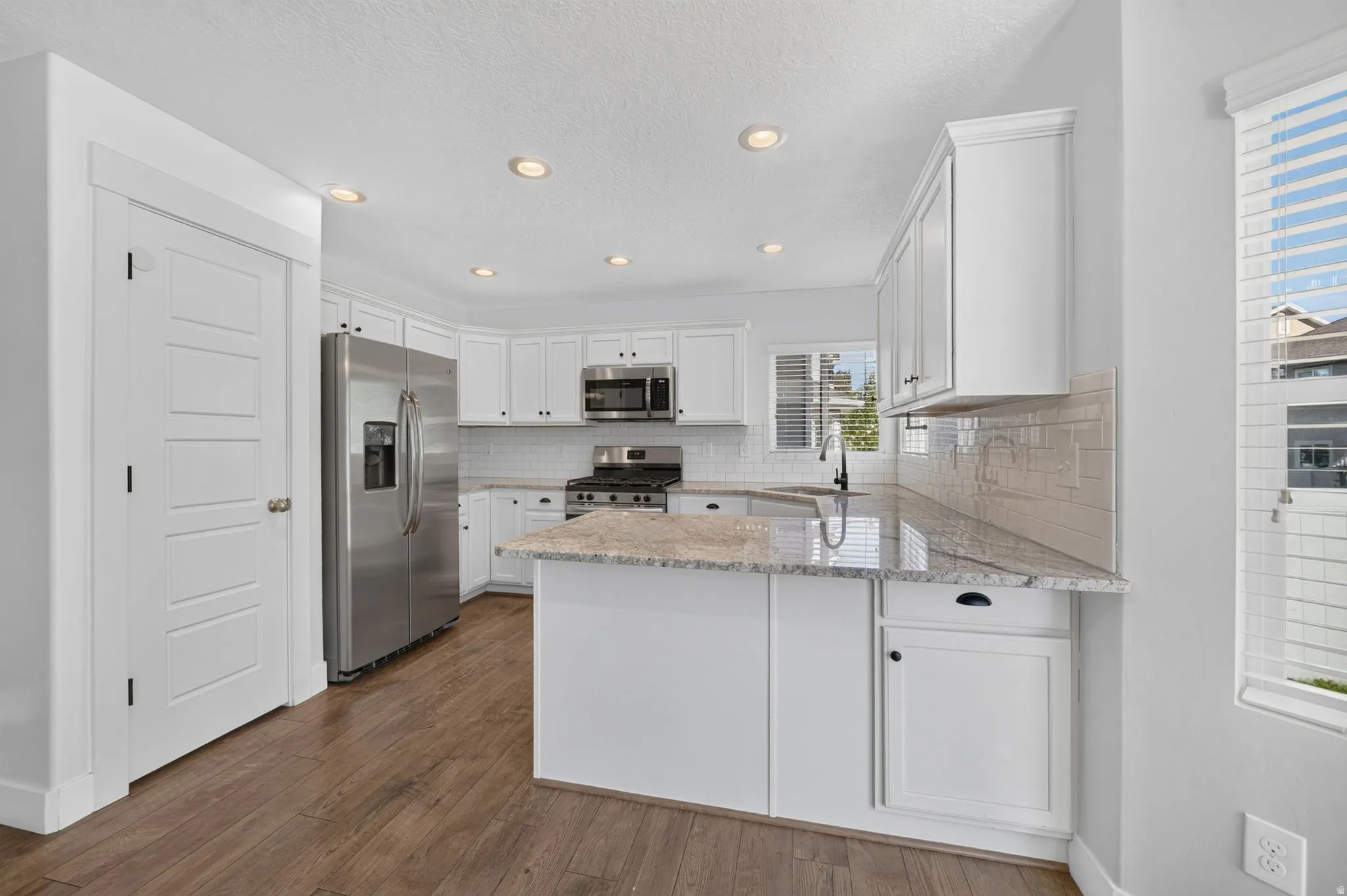 White shaker-style cabinetry with dark hardware, stainless steel appliances, light-toned stone countertops, subway tile backsplash, and wood-finish flooring