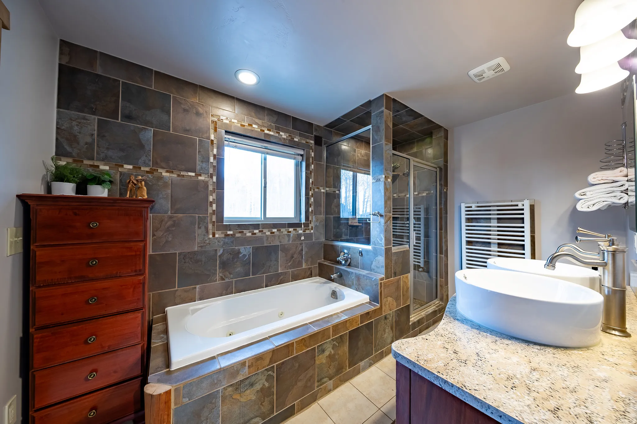 Tiled shower with glass enclosure, separate soaking tub, dual vessel sinks, and light-toned countertop
