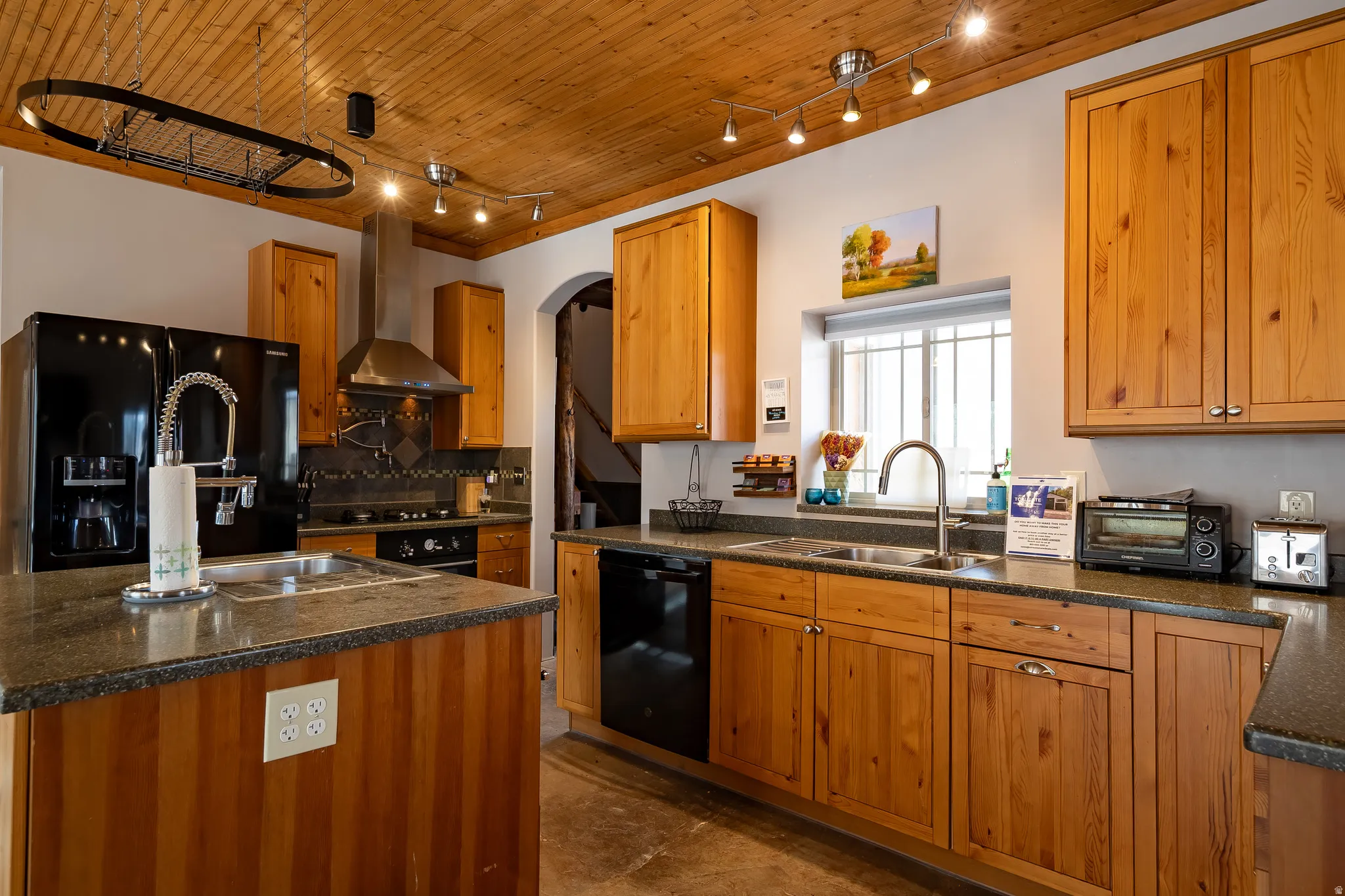 Wood-paneled ceiling with track lighting, wood cabinetry, dark countertops, black appliances, and a stainless steel range hood