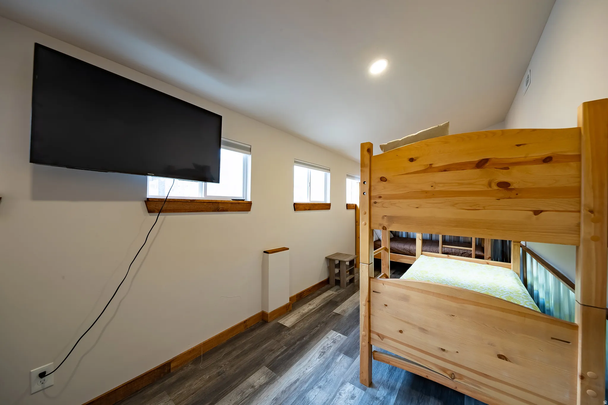 Wall-mounted television, wood-finish flooring, recessed ceiling lighting, natural wood bunk beds, and multiple windows