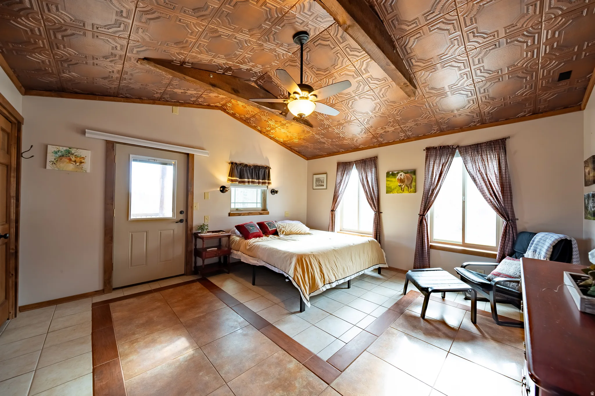 Vaulted ceiling with decorative tin-look panels, exposed wood beams, ceiling fan light fixture, multiple windows, and ceramic tile flooring with a wood-look border