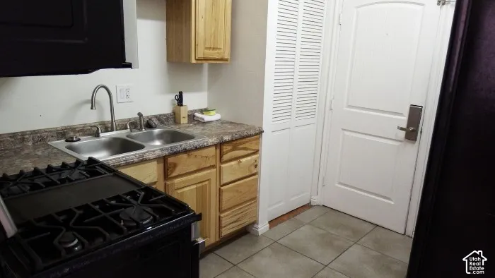 Black gas range, double basin stainless steel sink, wood-finish cabinetry, patterned countertop, and tiled flooring