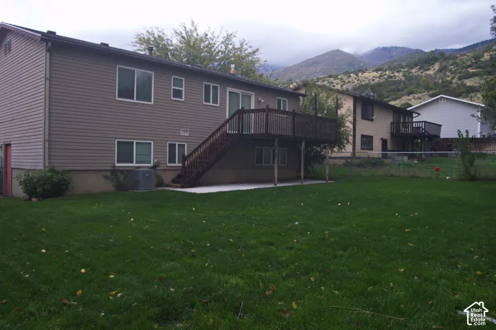 Exterior featuring vinyl siding, elevated wooden deck, concrete patio, expansive lawn, and mountain backdrop