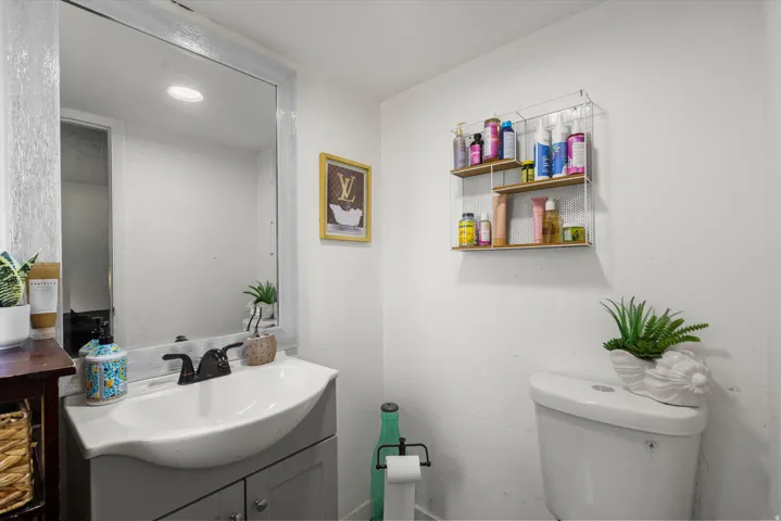 Vanity with an integrated sink and base cabinet, framed mirror, wall-mounted shelving unit, and a toilet with a top-mounted flush