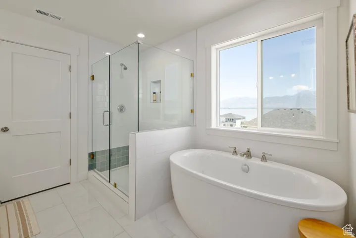 Freestanding soaking tub with a large window, glass-enclosed shower with tile surround, and tile flooring