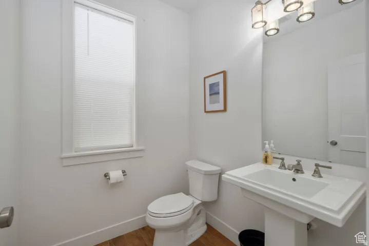 Pedestal sink with polished chrome hardware, white toilet, single window with blinds, wood-finish flooring, and wall-mounted light fixture