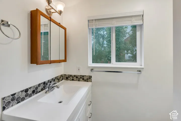 White vanity with a rectangular integrated sink, hexagonal mosaic tile backsplash, wood-finish medicine cabinet, polished chrome faucet, and a sliding window with window blinds