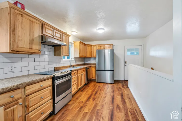 Kitchen featuring medium-tone wood cabinetry, stone-finish countertops, subway tile backsplash, stainless steel appliances, and wood-finish flooring