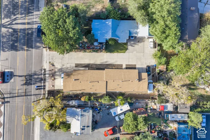 Aerial perspective featuring an asphalt shingle roof, extensive concrete parking surface, and mature trees on the lot