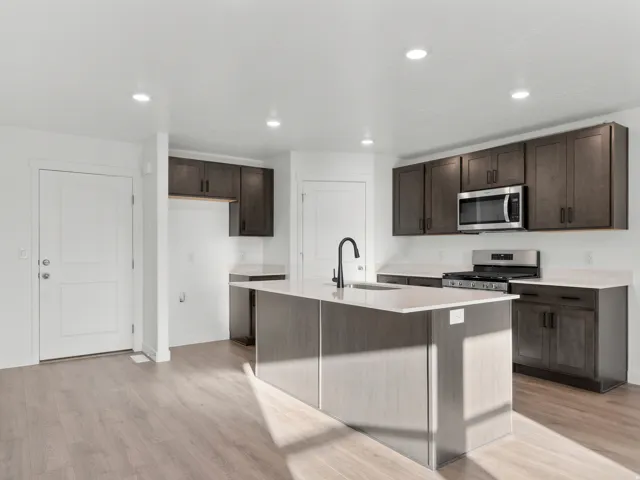 Modern kitchen featuring dark wood cabinetry, a large center island with waterfall-edge countertop, stainless steel appliances including a gas cooktop and microwave, pendant lighting, and wood-finish flooring throughout the open living space