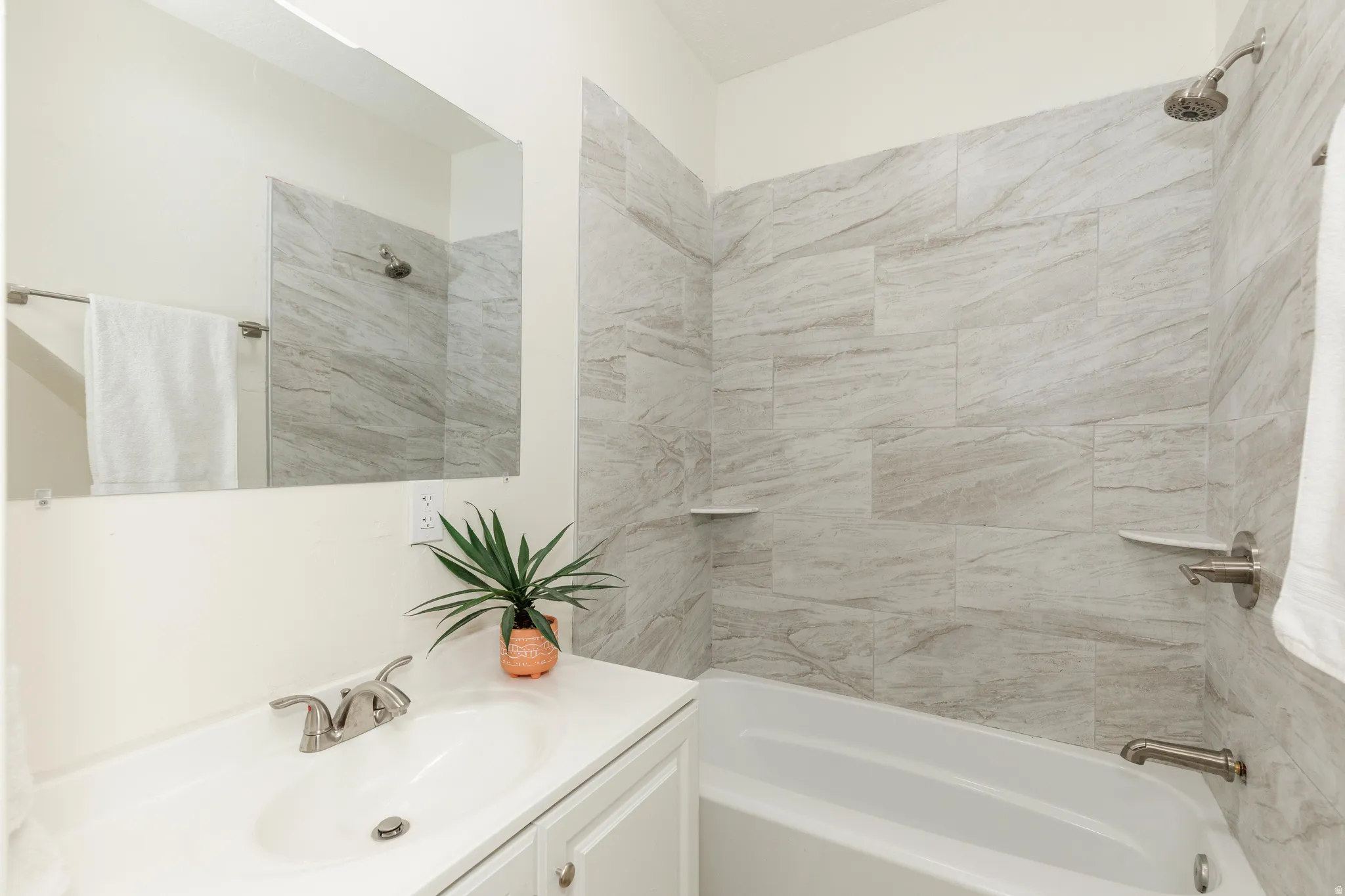 White vanity with an integrated sink and brushed nickel faucet, large wall mirror, and a tub-shower combination with light-toned tiled walls