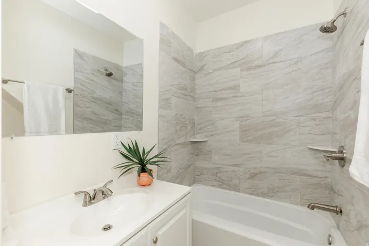 White vanity with an integrated sink and brushed nickel faucet, large wall mirror, and a tub-shower combination with light-toned tiled walls