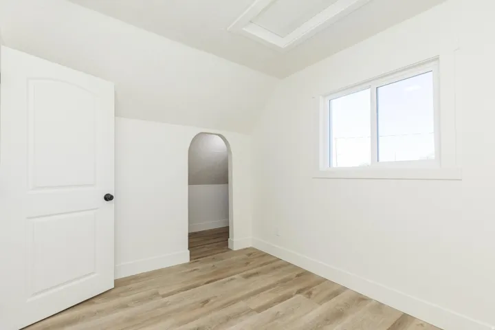 Wood-finish flooring, white paneled door with black hardware, white walls, window with white trim, and arched alcove