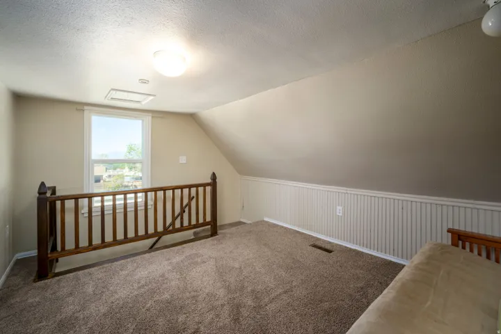 Sloped ceiling room featuring a natural wood-finish railing, single window, and textured carpet flooring