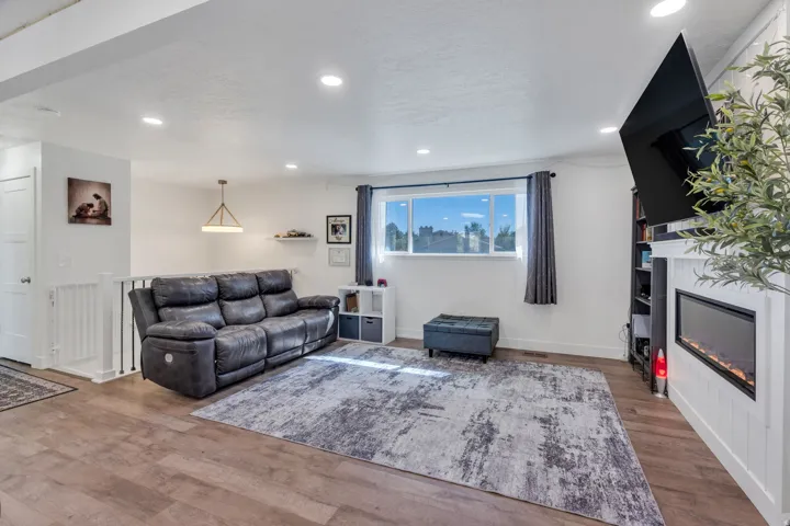Recessed lighting, wood-finish flooring, white paneled fireplace, large window with dark drapes, and a linear electric fireplace insert