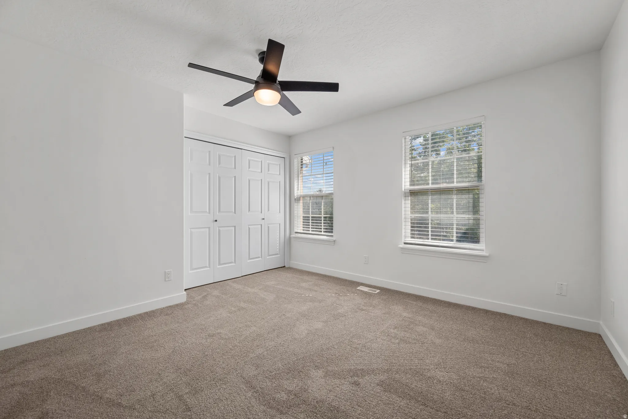 Neutral carpeting, two windows with blinds, a ceiling fan with integrated lighting, and a closet with bifold doors