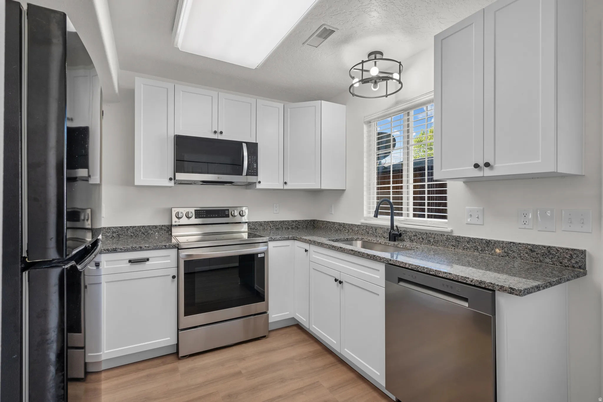 White shaker-style cabinetry, stainless steel appliances, granite countertops, wood-finish flooring, and a modern ceiling light fixture