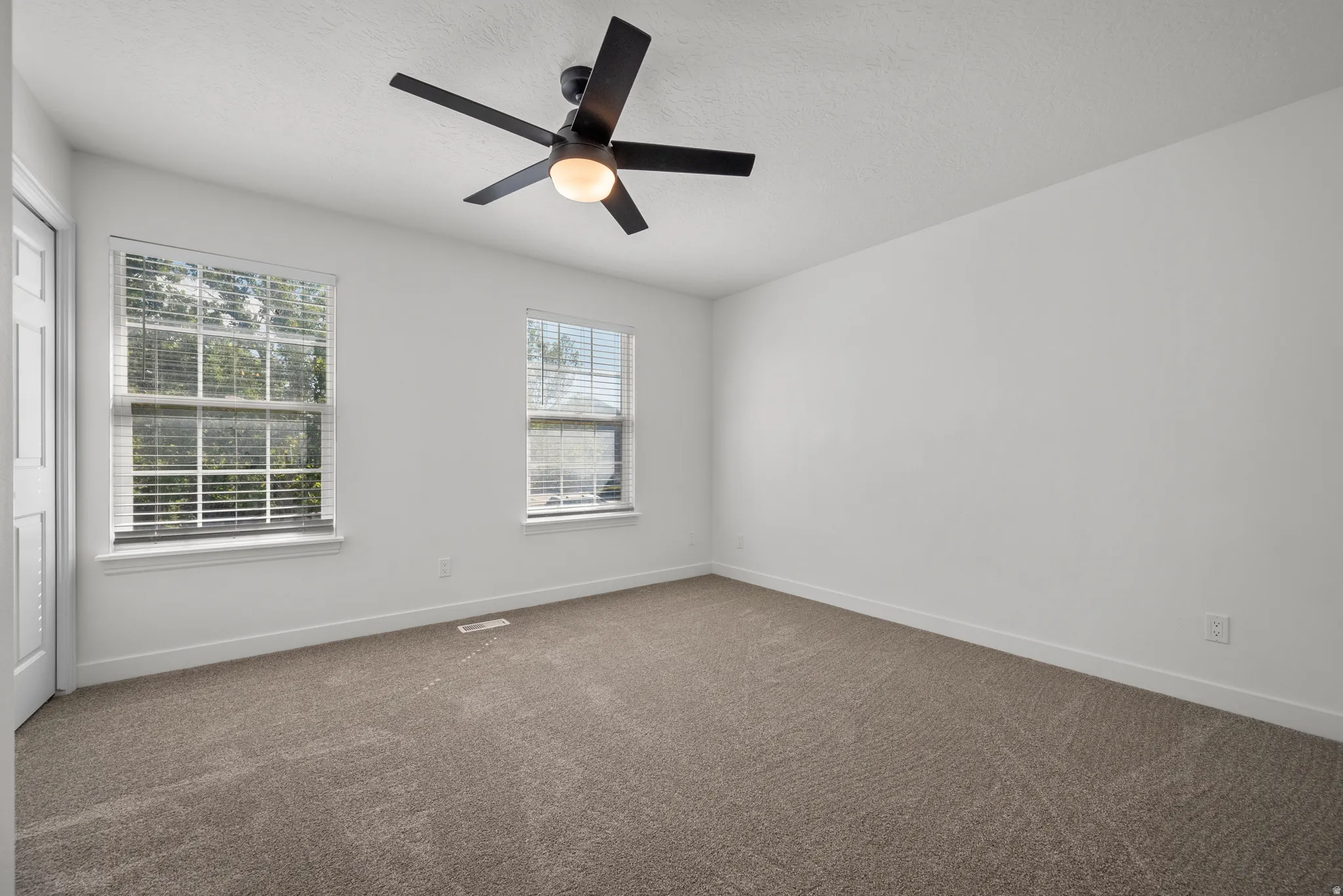 Neutral carpet flooring, white walls, two windows with blinds, ceiling fan with integrated lighting, and a white door