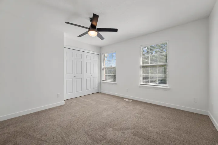 Neutral carpeting, two windows with blinds, a ceiling fan with integrated lighting, and a closet with bifold doors
