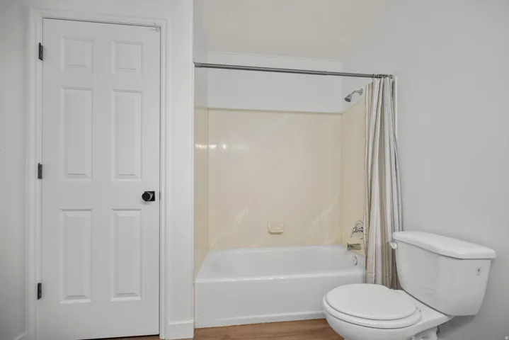 White paneled door with black hardware, white toilet, white bathtub, and wood-finish flooring