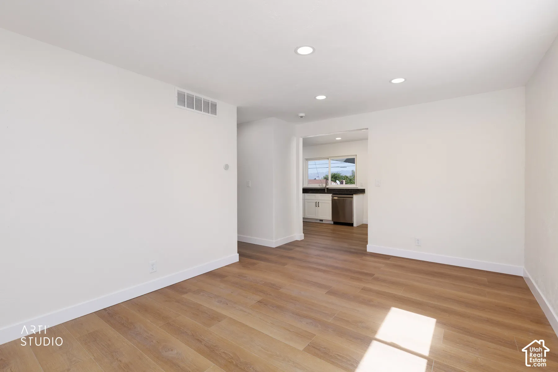 Wood-finish flooring, recessed lighting, white walls, and a separate kitchen area with white cabinetry and a stainless steel dishwasher