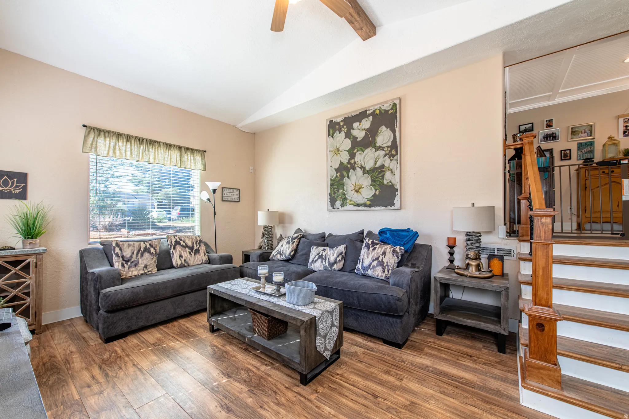 Vaulted ceiling with exposed wood beam, wood-finish flooring, staircase with wood banister and white risers, large window with blinds, and built-in shelving unit