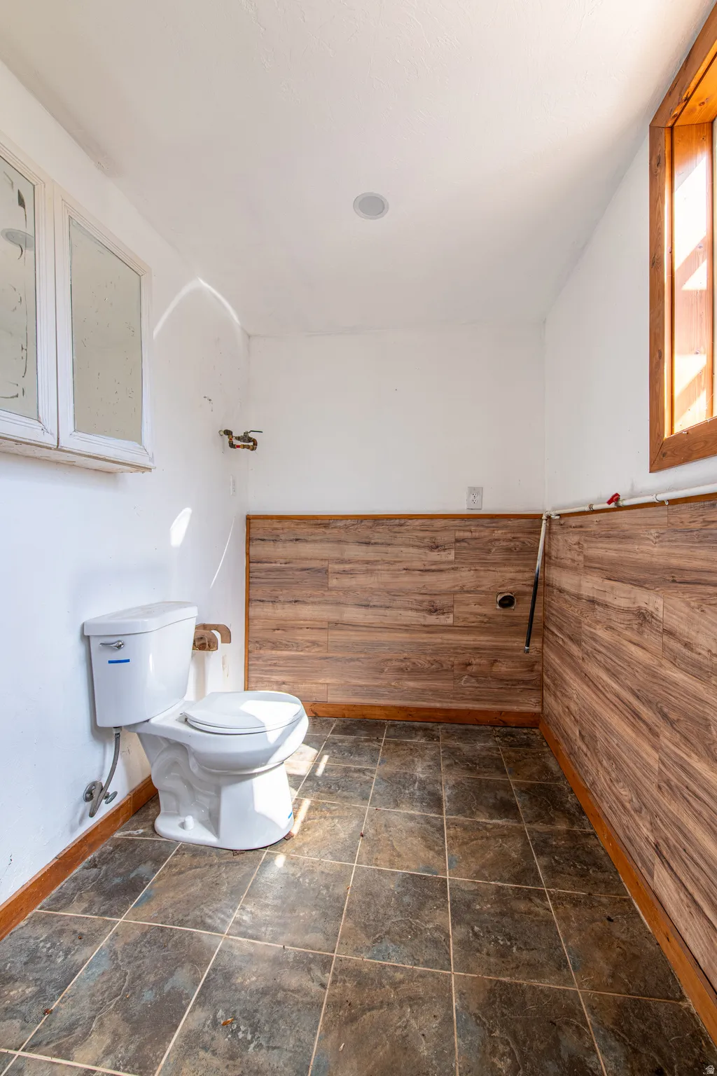 White toilet with exposed plumbing, wood-finish wainscoting, and large format floor tiling