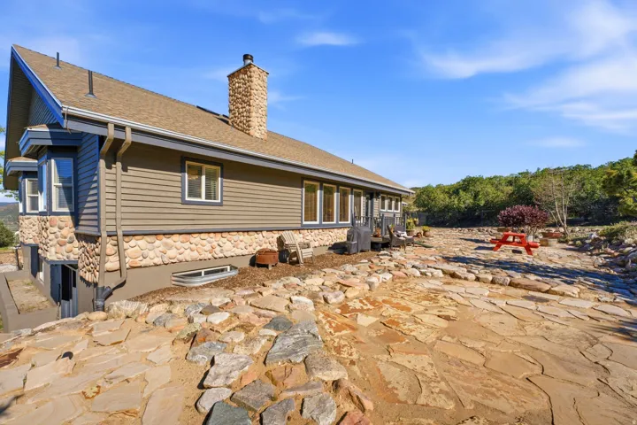 Stone and siding exterior with a prominent stone chimney, multiple windows, and a large flagstone patio