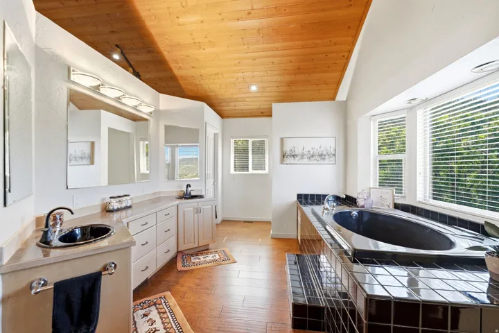 Wood-paneled ceiling, dual vanities with integrated sinks, built-in soaking tub with dark tile surround, and multiple windows