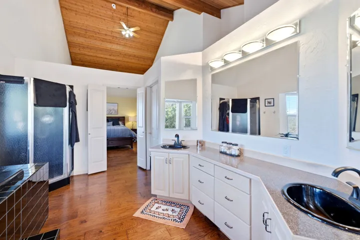 Vaulted wood-paneled ceiling with ceiling fan, dual vanity with integrated sinks, wood-finish flooring, and a tiled shower enclosure