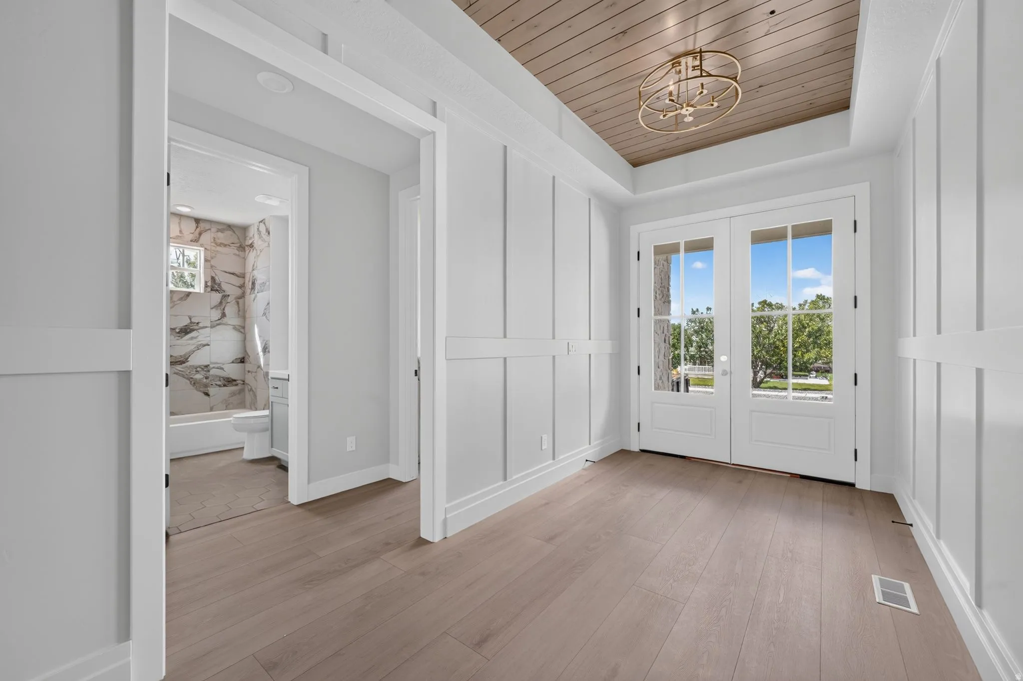 Wood-finish flooring, white paneling, wood-paneled ceiling, double glass-paneled doors, and a gold-tone chandelier