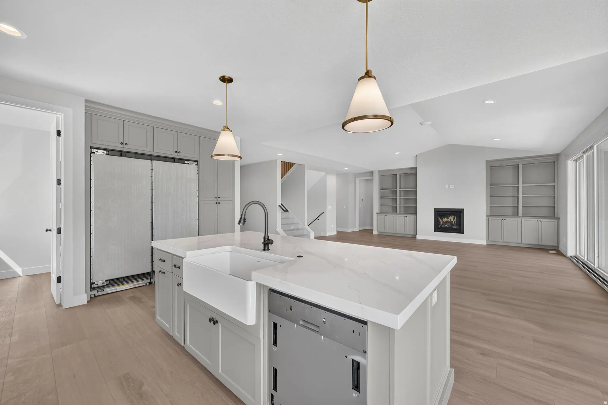 Kitchen island with white countertop and farmhouse sink, gray cabinetry, wood-finish flooring, vaulted ceiling, and built-in shelving with fireplace