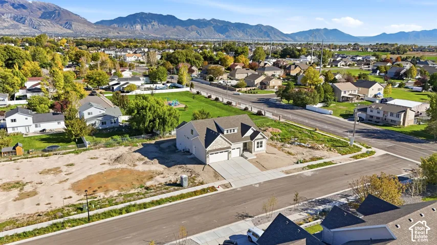 Aerial perspective featuring a multi-story residence with a multi-car garage, shingled roof, mountain backdrop, and expansive corner lot