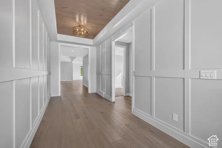 Hallway featuring wood-finish flooring, floor-to-ceiling board and batten wainscoting, recessed tray ceiling with wood-grain inlay, and a metallic drum chandelier