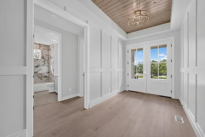 Wood-finish flooring, white paneling, wood-paneled ceiling, double glass-paneled doors, and a gold-tone chandelier