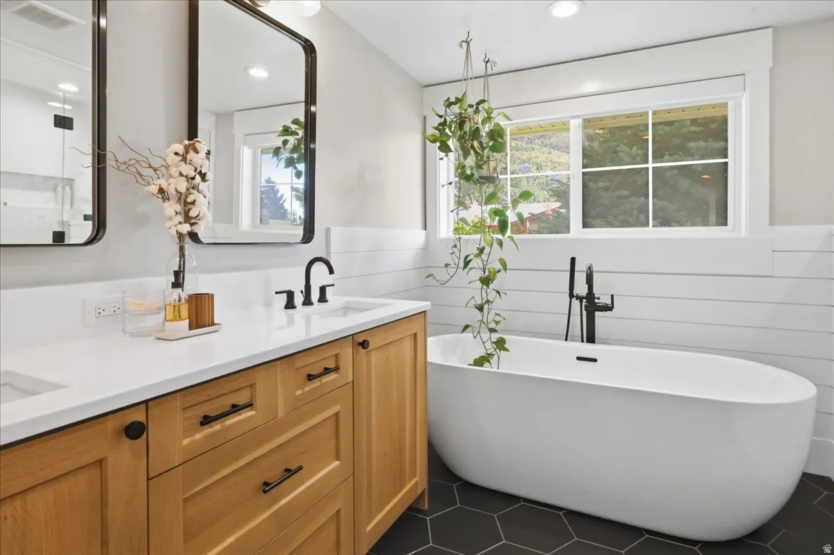 Freestanding oval soaking tub with matte black fixtures, dual vanity with wood cabinetry, crisp white countertop, and hexagon tile flooring