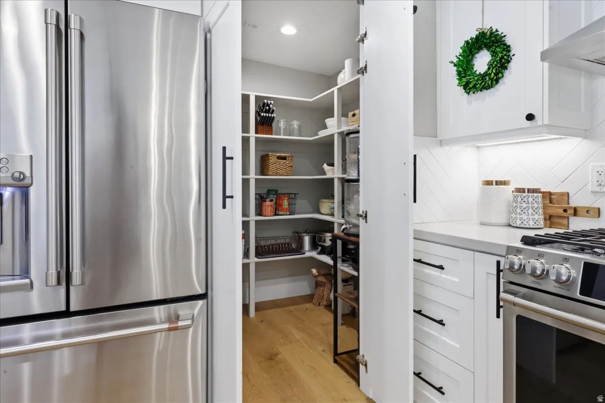 Stainless steel refrigerator with a water dispenser, white shaker-style cabinetry with black hardware, gas range, light wood-finish flooring, and a white countertop