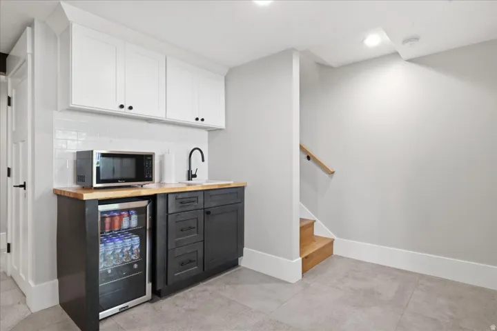 Wet bar featuring white upper cabinetry, dark lower cabinetry, a wood-finish countertop, a stainless steel beverage refrigerator, and a matte black faucet