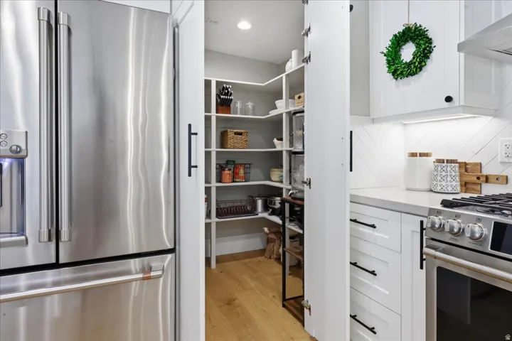 Stainless steel refrigerator with a water dispenser, white shaker-style cabinetry with black hardware, gas range, light wood-finish flooring, and a white countertop