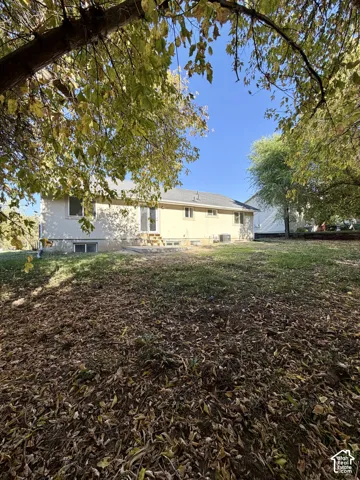 Rear exterior featuring a neutral-toned stucco finish, a central concrete patio, multiple windows, and mature trees
