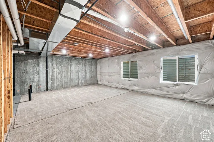 Unfinished basement featuring exposed wood joists, concrete foundation walls, two windows, and gray patterned carpeting