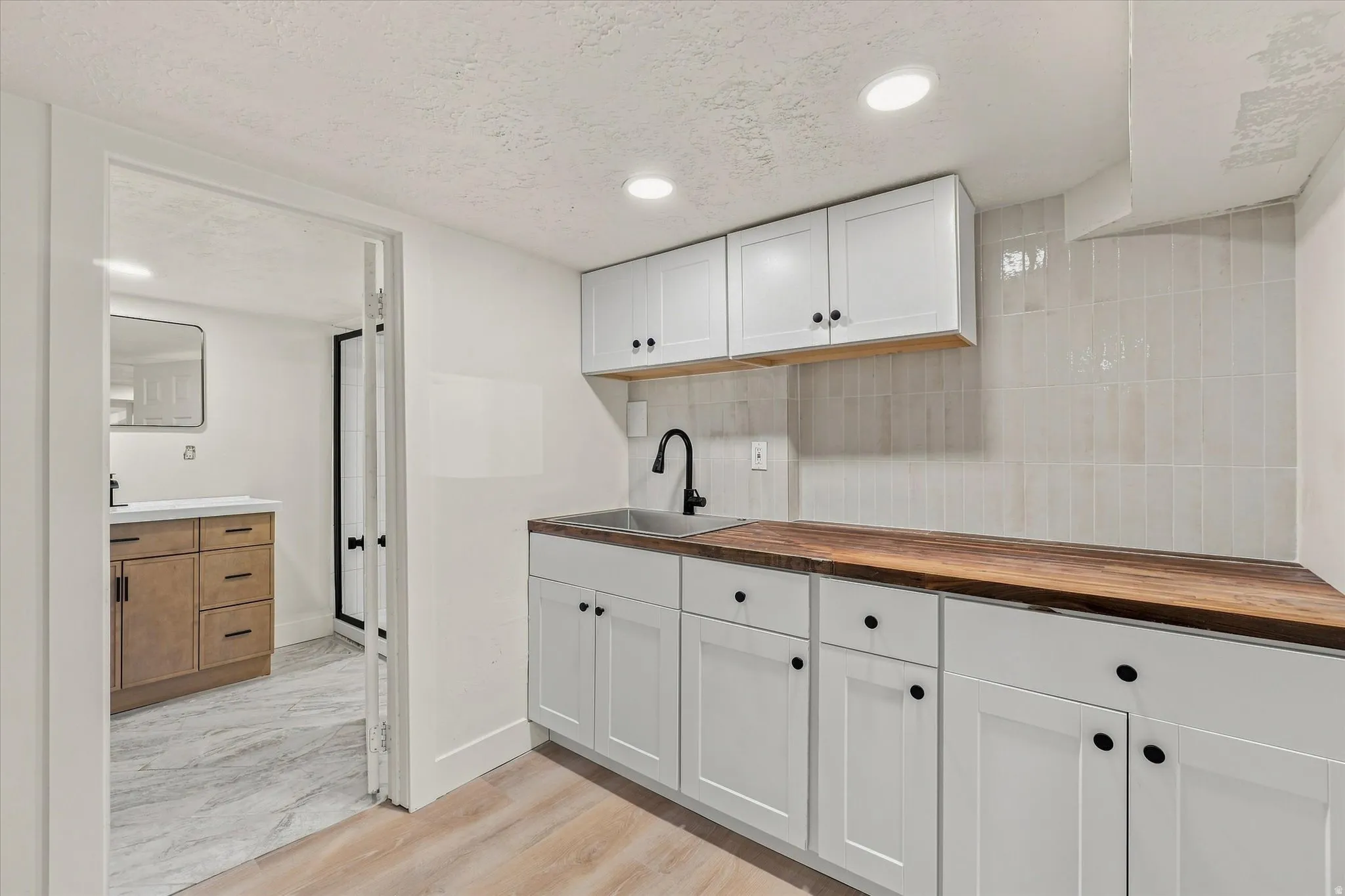 White shaker cabinetry with black hardware, wood-finish countertops, stainless steel sink with matte black faucet, and vertical tile backsplash