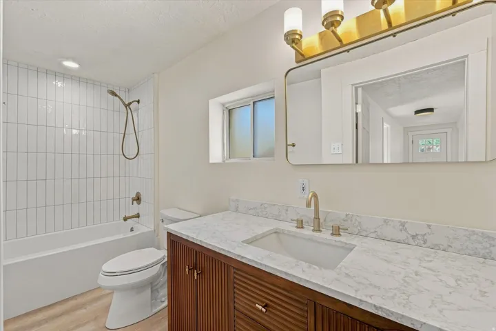 White subway tile tub surround, wood-finish vanity with fluted details, rectangular undermount sink, stone-look countertop, and gold-finish fixtures