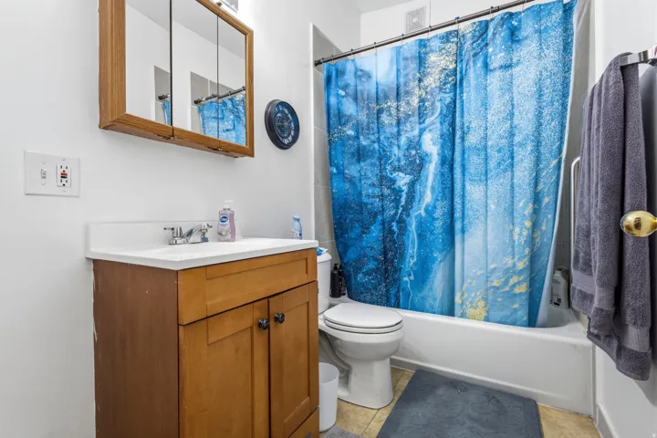 White ceramic tile flooring, wood-finish vanity with integrated sink, chrome faucet, wall-mounted mirrored medicine cabinet, and white bathtub with shower
