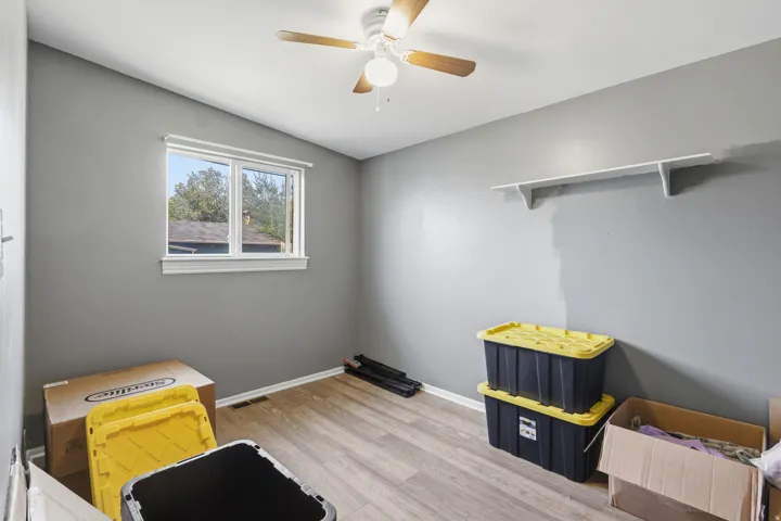 Wood-finish flooring, neutral gray wall tones, ceiling fan with light fixture, single window with white trim, wall-mounted shelving